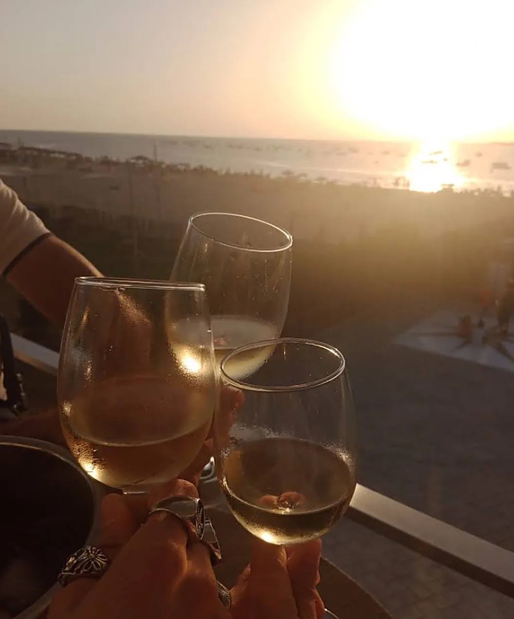 Dos manos brindando con copas de vino blanco frente a la playa al atardecer