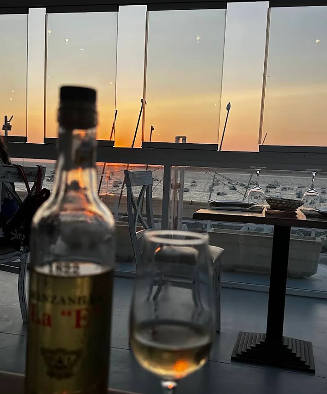 Botella de Manzanilla La E y una copa en la mesa, con el atardecer naranja sobre el río detrás del cristal