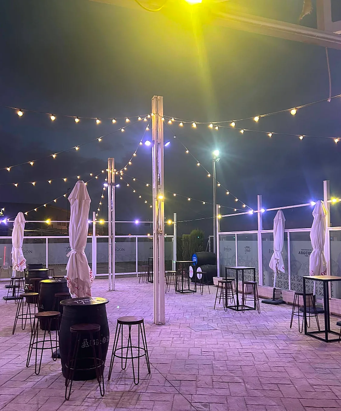 Terraza de El Espigón de noche, con guirnaldas de bombillas, mesas altas de barril y sombrillas cerradas